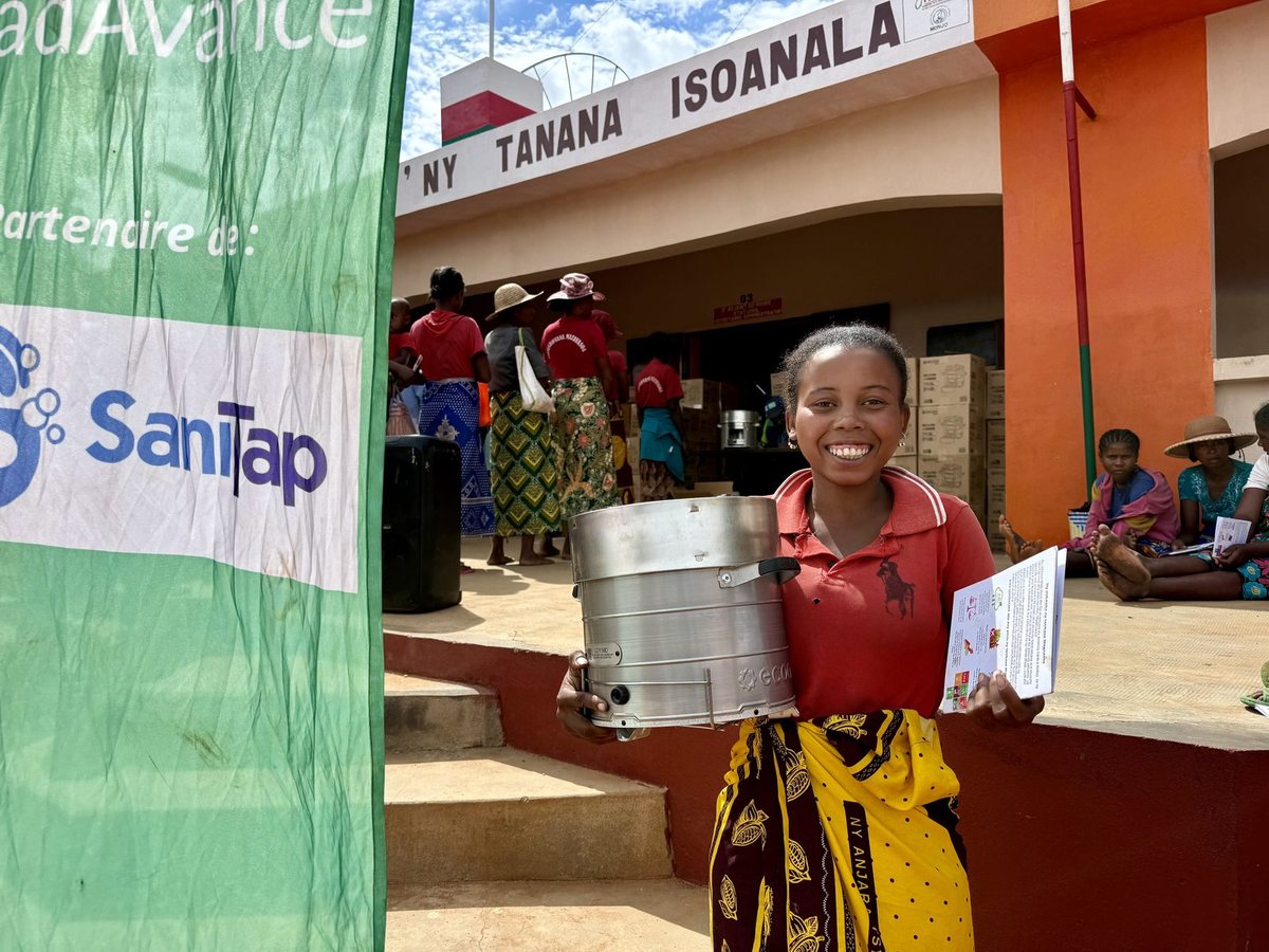 A smiling young woman holds a new stainless steel cookstove and a leaflet at a stove distribution beside a SaniTap banner.