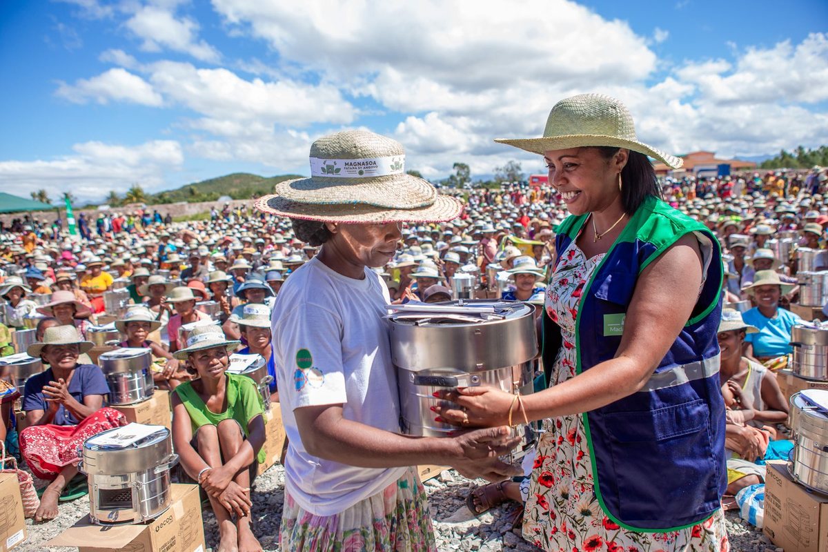 A smiling woman in a SaniTap vest hands a stainless steel cookstove to a woman in a straw hat before a large seated crowd at the launch.