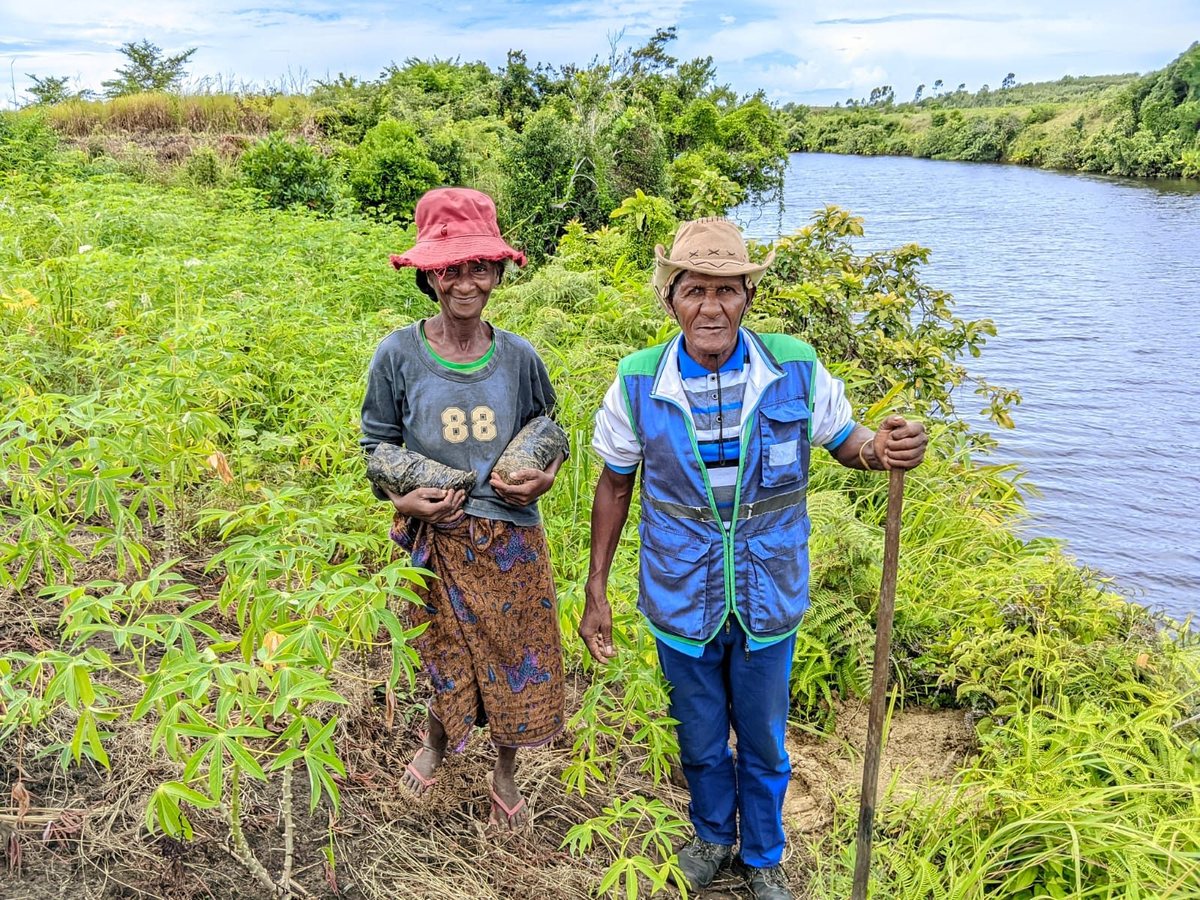 Two smiling older villagers, one holding bamboo seedlings in bags and the other a stick, stand in green vegetation beside a river.
