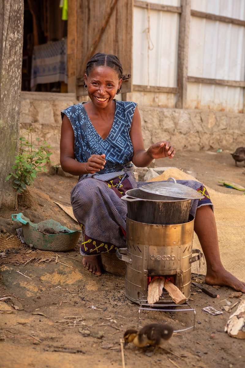 A woman laughs while sitting beside a lit improved cookstove with a pot and firewood, as a duckling pecks at the ground in the foreground.